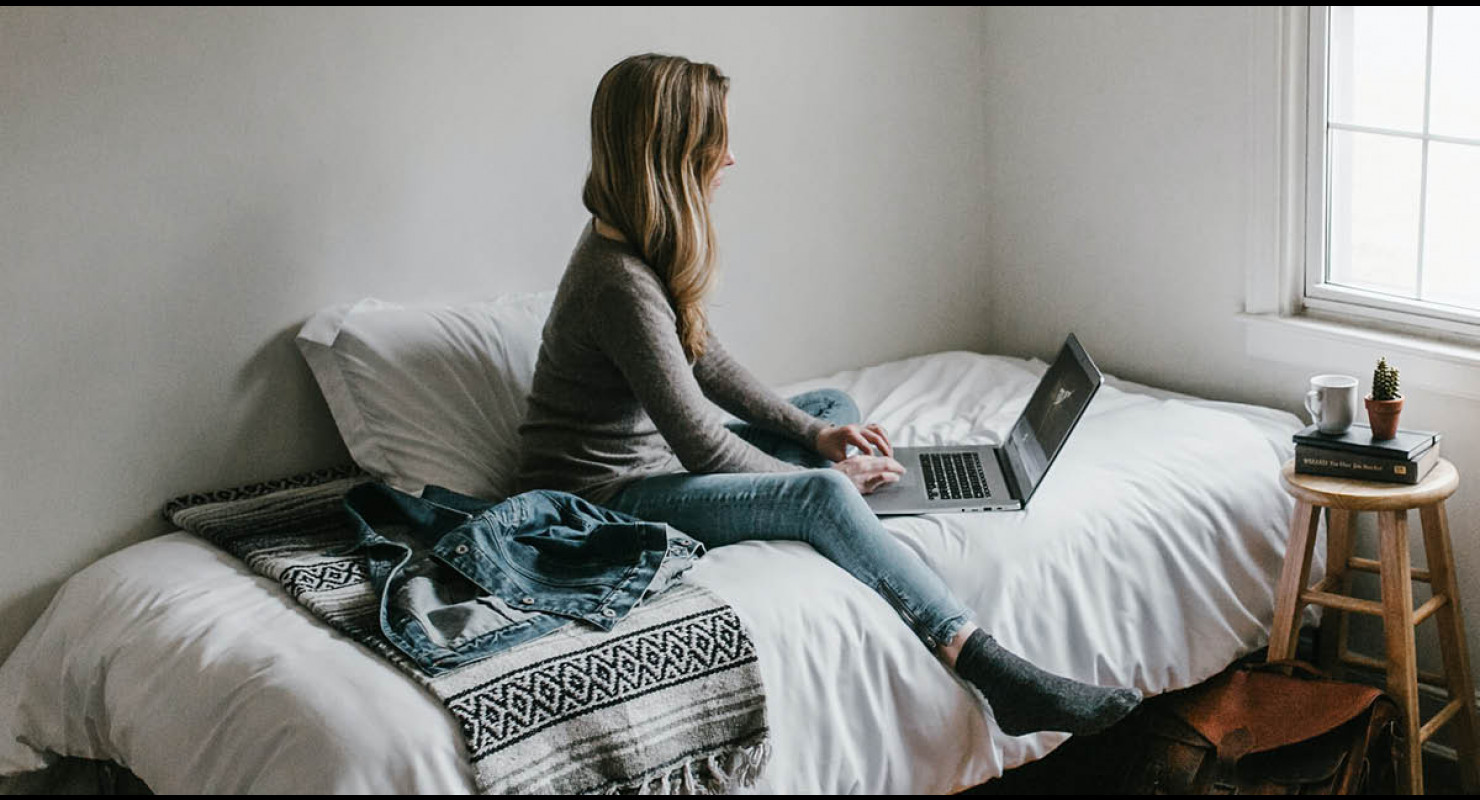 Étudiante en train de faire ses recherches sur son ordinateur, assise sur son lit dans son appartement